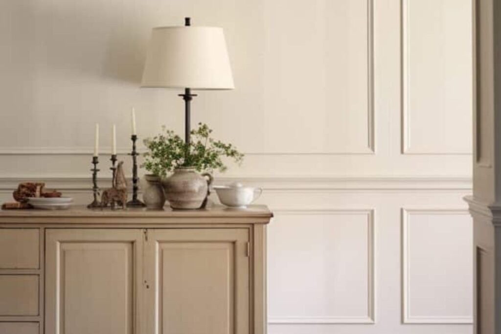 A soft beige cabinet with a table lamp, small plants, and candles, set against white walls white trim.