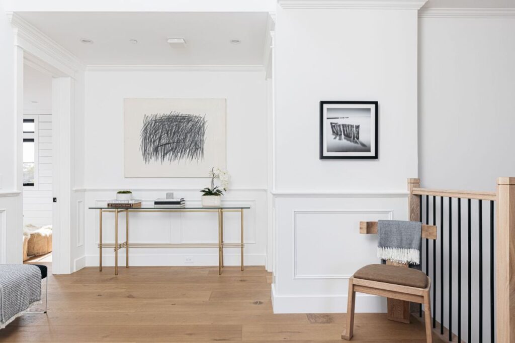 A minimalistic hallway with a glass table, artwork, and a black-and-white photograph, with white walls white trim and a wooden floor.