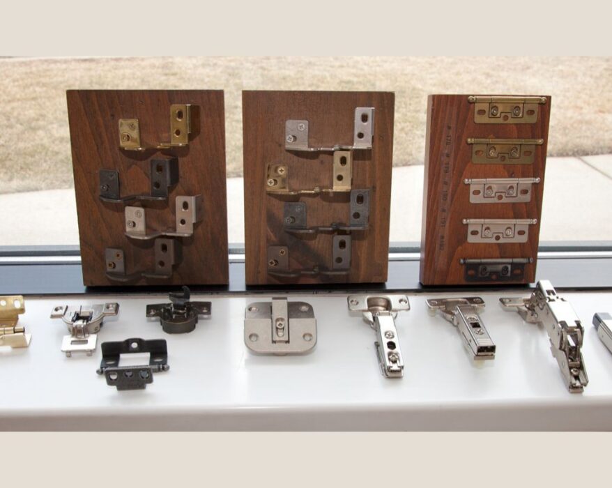 Various types of hidden cabinet hinges displayed on wooden boards, showcasing different designs and finishes. The hinges are arranged for easy comparison, including several metal finishes and mechanisms that are typically used for modern, concealed cabinetry solutions.