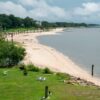 A scenic view of a beach in Ocean Springs, MS, showcasing things to do in the area. The beach features soft, sandy shores stretching into the distance, bordered by green grass and a few white chairs set up near the water’s edge. Trees and a pier are visible in the background, and the sky is partly cloudy. The water is calm and smooth, creating a peaceful and tranquil atmosphere, perfect for relaxation and exploring the things to do in Ocean Springs MS.