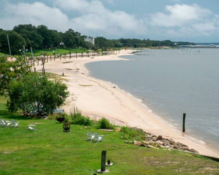 A scenic view of a beach in Ocean Springs, MS, showcasing things to do in the area. The beach features soft, sandy shores stretching into the distance, bordered by green grass and a few white chairs set up near the water’s edge. Trees and a pier are visible in the background, and the sky is partly cloudy. The water is calm and smooth, creating a peaceful and tranquil atmosphere, perfect for relaxation and exploring the things to do in Ocean Springs MS.