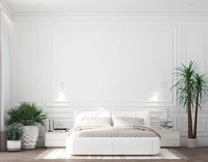 A modern bedroom featuring a clean, minimalistic design with white walls and white trim. The room includes a white bed with soft beige linens, a cozy rug, and decorative plants. On the side, there's a nightstand with a lamp and a small decorative sailboat. Large windows with sheer curtains allow natural light to fill the room, emphasizing the bright, airy atmosphere created by the white walls and white trim.