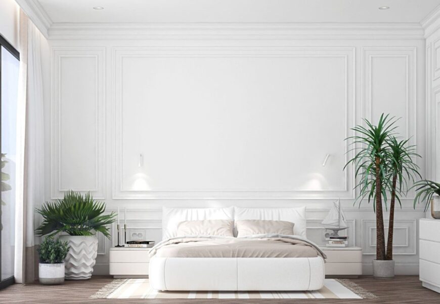 A modern bedroom featuring a clean, minimalistic design with white walls and white trim. The room includes a white bed with soft beige linens, a cozy rug, and decorative plants. On the side, there's a nightstand with a lamp and a small decorative sailboat. Large windows with sheer curtains allow natural light to fill the room, emphasizing the bright, airy atmosphere created by the white walls and white trim.