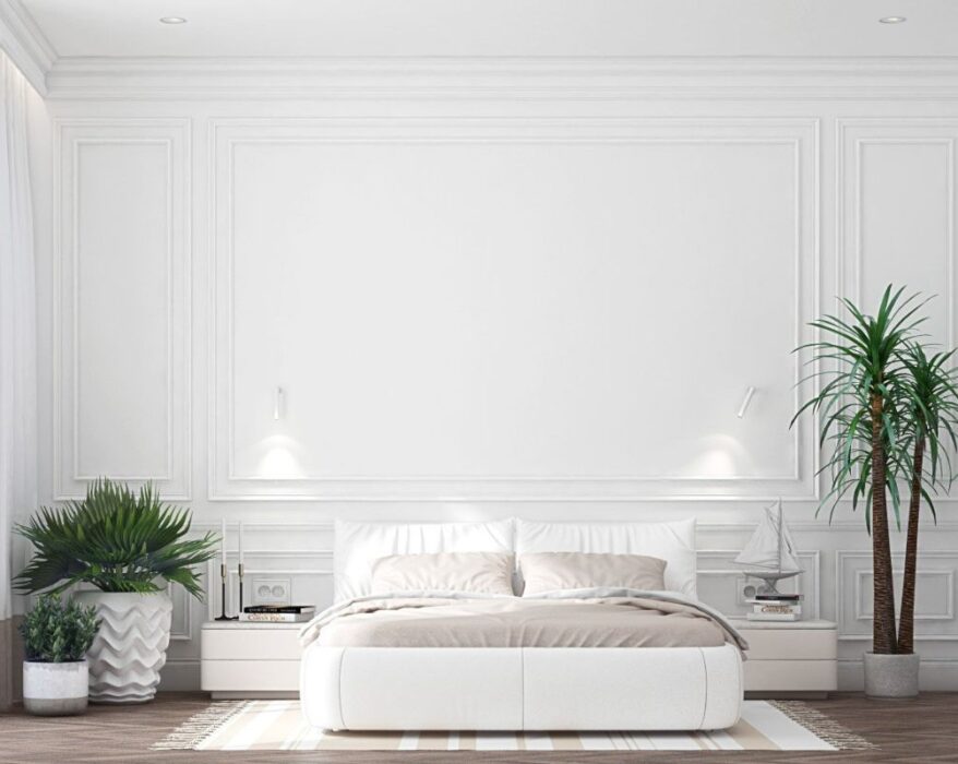 A modern bedroom featuring a clean, minimalistic design with white walls and white trim. The room includes a white bed with soft beige linens, a cozy rug, and decorative plants. On the side, there's a nightstand with a lamp and a small decorative sailboat. Large windows with sheer curtains allow natural light to fill the room, emphasizing the bright, airy atmosphere created by the white walls and white trim.